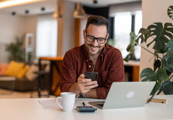 Attractive man looking at his phone with laptop in front of him, doing work early in the morning