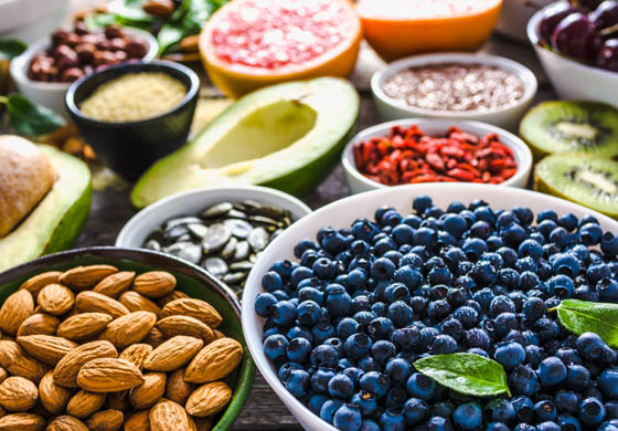 Bowls of nutritious foods including blueberries, almonds, avocado, pumpkin seeds, citrus, kiwis, and more