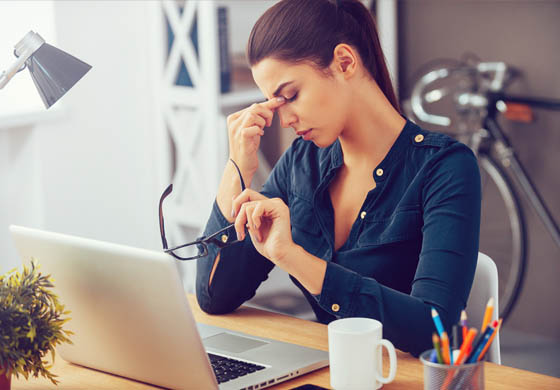 Woman stressed out at her desk, with hand holding the bridge of her nose and her glasses in the other hand