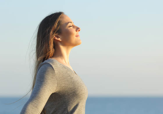 Beautiful woman with her eyes closed, breathing deeply at the seaside with sun rising behind her